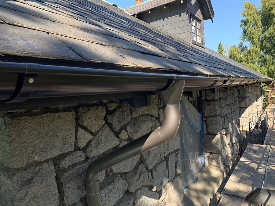 A dark brown round Lindab Rainline gutter attached to the shingled roof of a house with a rough stone facade.