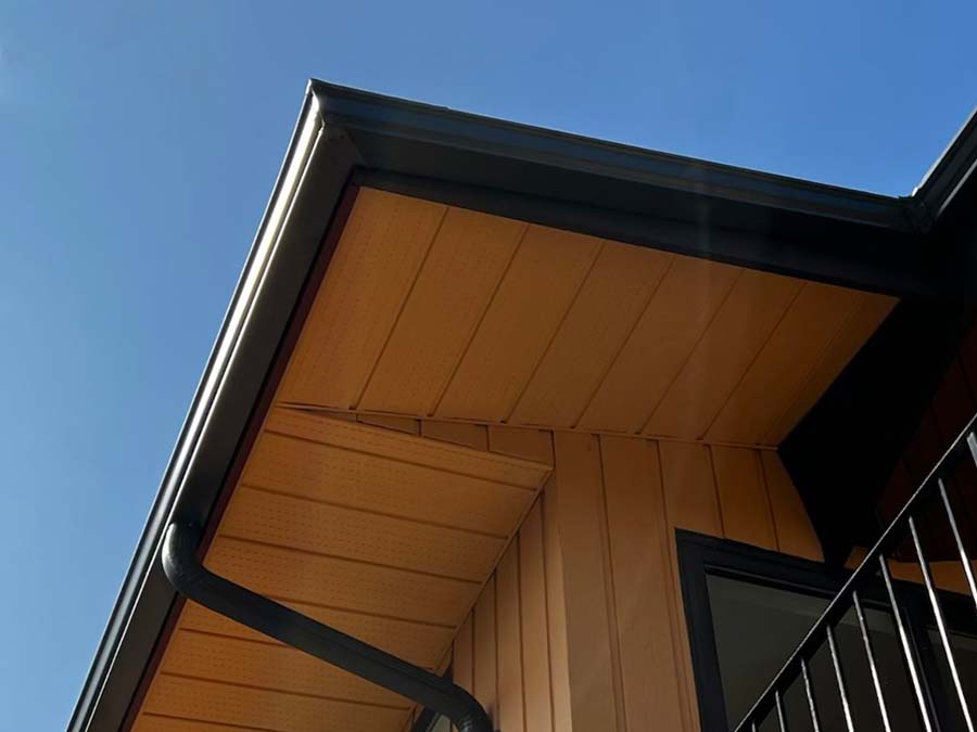 An upwards shot of a black aluminum continuous gutter and downspout, installed on a house with wood-coloured soffit and walls, and black trim.
